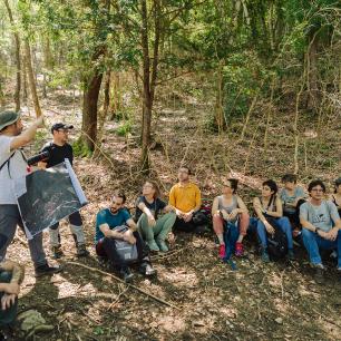 Group of people sitting in a forest, listening to a presentation.