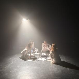 Four people dressed in white sitting on the floor among papers under the light of a spotlight 