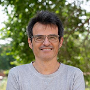 Smiling man with glasses, outdoors, in front of greenery.
