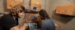 Two men looking at laptop near wooden boxes and control panel. CREAF
