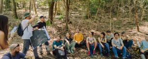 Group of people sitting in a forest, listening to a presentation.