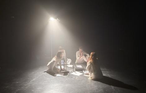 Quatre persones vestides de blanc, assegudes a terra enmig de piles de papers sota la llum d'un focus. 