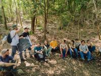 Group of people sitting in a forest, listening to a presentation.