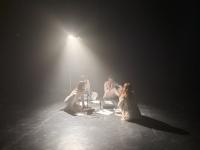 Four people dressed in white sitting on the floor among papers under the light of a spotlight 