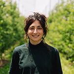 Smiling woman, dark hair, wearing green turtleneck, background of green plants.