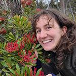 Woman smiles, posing near red flowers and green plants.