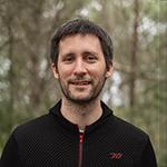Smiling man in black sweater, forest background.