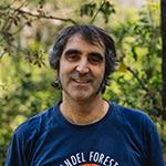 Man smiling, wearing a blue shirt, standing outside with trees in the background.