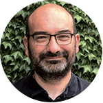 Man with glasses smiling in front of green leaves.