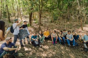 Group of people sitting in a forest, listening to a presentation.