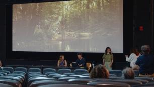 People on a stage with a screen displaying a forest scene in a movie theater.