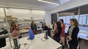 Group of people in a lab, conversing, near equipment and a window.